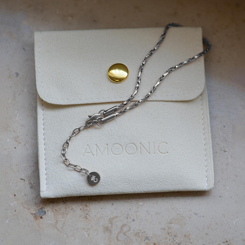Eine elegante silberne Kette liegt auf einer hochwertigen, cremefarbenen Schmucktasche mit goldfarbenem Knopf und Amoonic-Prägung – luxuriöse Schmuckpräsentation.