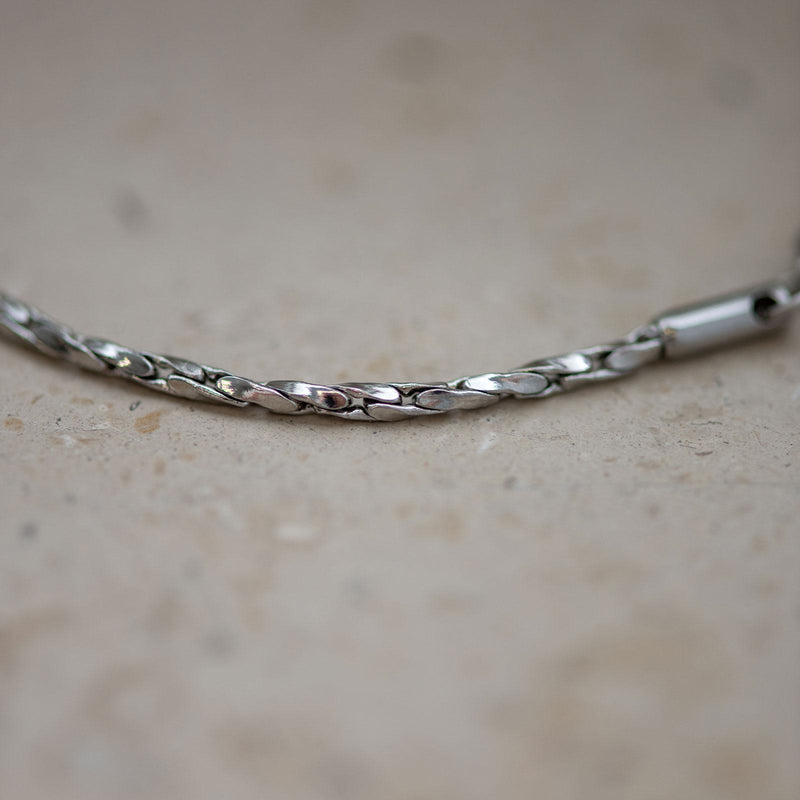 Makroaufnahme einer feingliedrigen Silberkette mit gedrehten, spiegelglatt polierten Gliedern. Die reflektierende Oberfläche fängt das Licht sanft ein und zeigt subtile Farbnuancen in silber und platin. Der Hintergrund besteht aus hellem, leicht strukturiertem Stein, der den Glanz der Kette elegant betont.