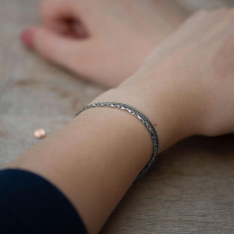 Ein zartes Silberarmband liegt eng am Handgelenk einer Person an. Die fein geschwungene Gliederstruktur reflektiert das Licht dezent und zeigt eine polierte, leicht gedrehte Oberfläche. Das Metall schimmert in kühlem Silberton, kontrastiert sanft mit der hellen Haut und dem matten Holzuntergrund im Hintergrund.