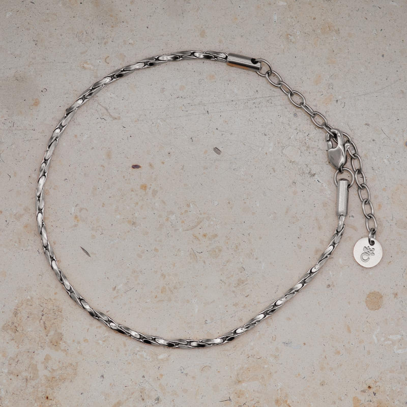 Ein filigranes Silberarmband liegt in ovaler Form auf hellem, leicht strukturiertem Steinuntergrund. Die Glieder der Kette sind sanft gedreht und fein poliert, wodurch sie das Licht mit einem subtilen, spiegelnden Glanz reflektieren. Am Ende befindet sich eine zarte Verlängerungskette mit rundem Anhänger, der das Amoonic-Logo trägt, sowie ein hochwertiger Karabinerverschluss in glänzendem Silberton.