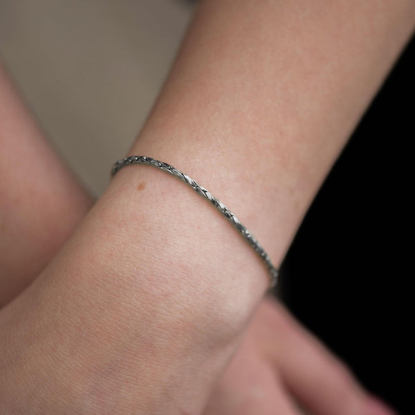 Ein schmales, glänzendes Silberarmband liegt elegant am Handgelenk einer Person. Die feinen, gleichmäßig gearbeiteten Glieder zeigen eine subtile Drehung, die das Licht weich reflektiert. Die helle Haut und der dunkle Hintergrund erzeugen einen klaren, stilvollen Kontrast, der die Struktur der Kette betont.